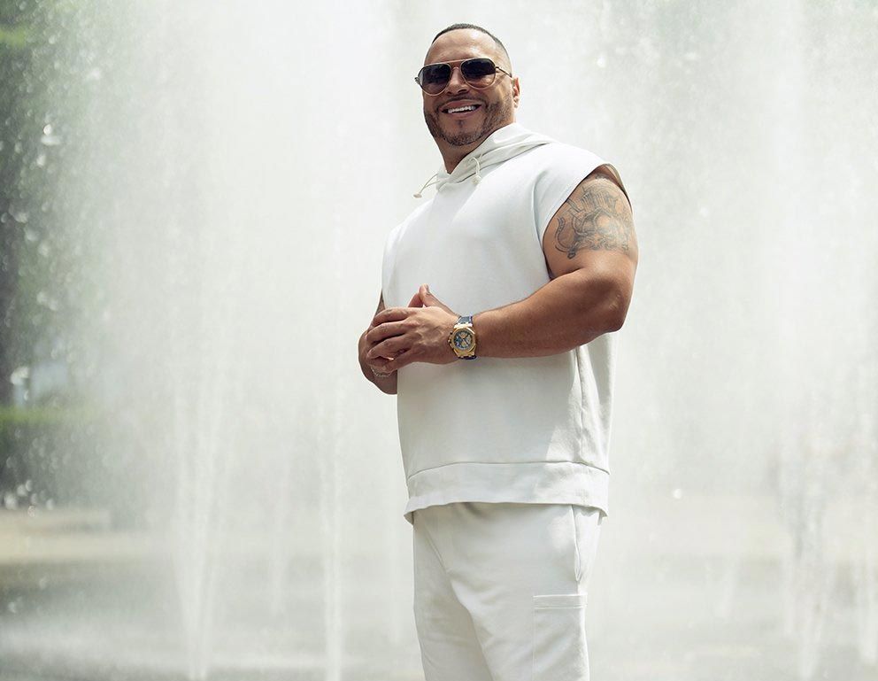 Man in white outfit stands in front of a fountain, smiling with hands clasped.