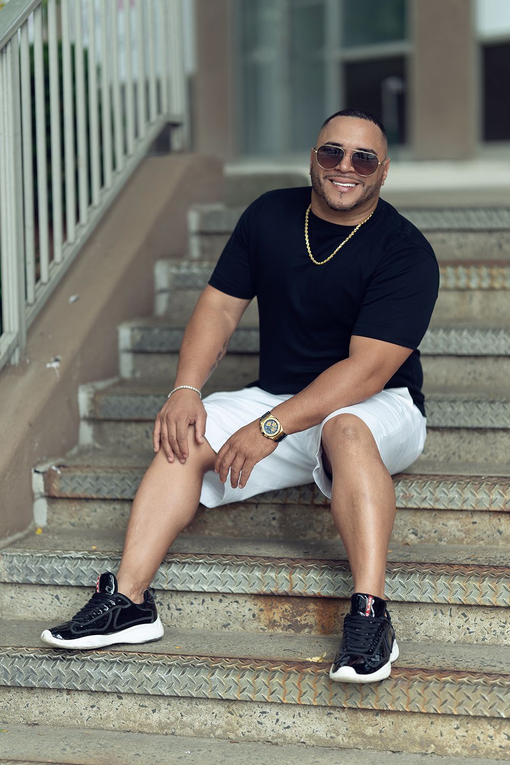 Man in black shirt, white shorts, and sunglasses sits on outdoor concrete stairs, smiling.