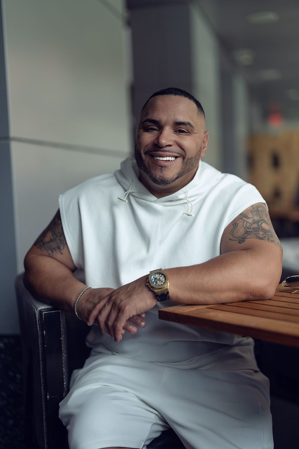 Man with tattoos in white sleeveless hoodie smiles, resting on a wooden table.