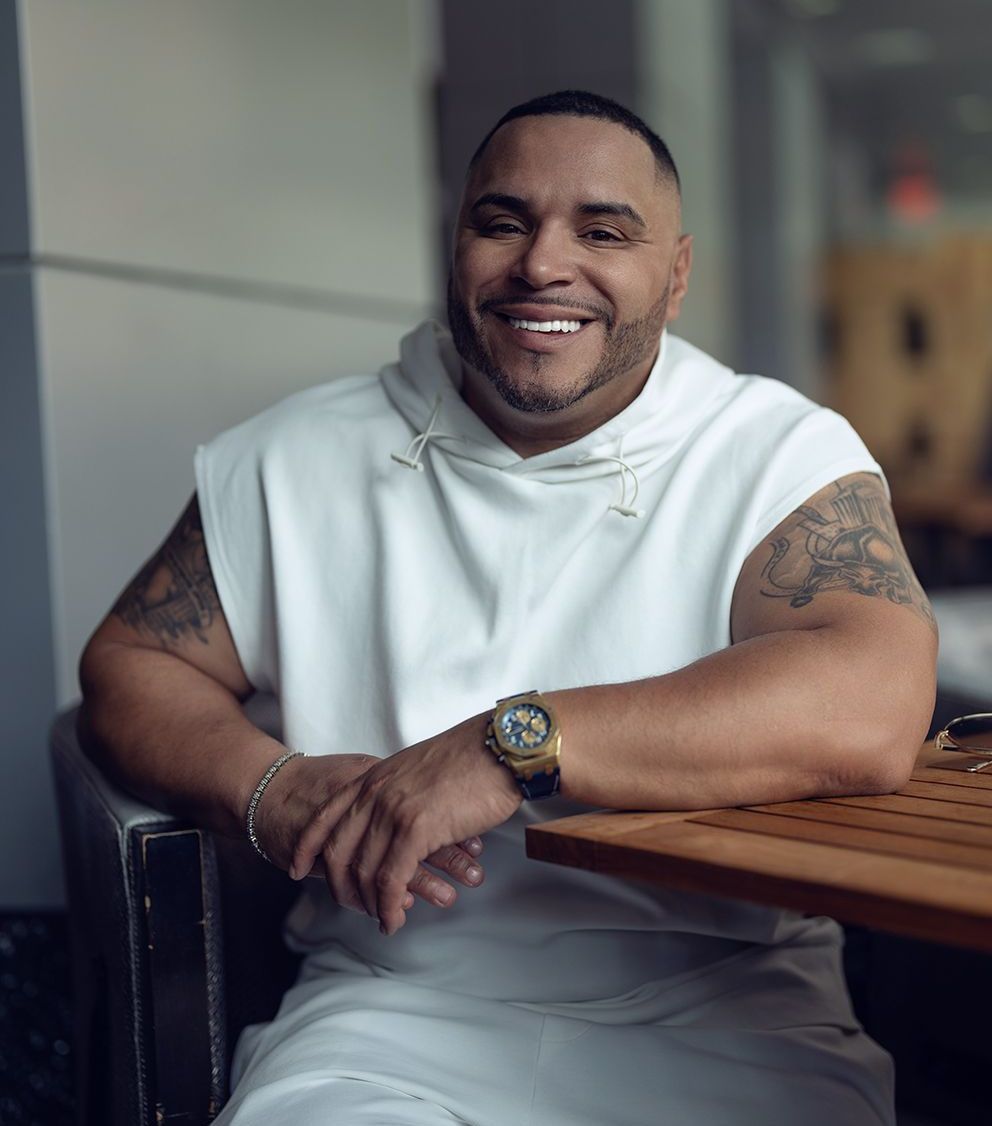 Man in white sleeveless hoodie smiles, resting arm on a table; visible arm tattoos.