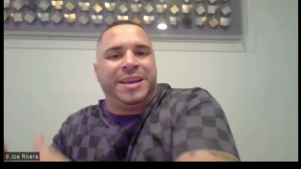 Man in checkered shirt, speaking into a webcam with hand gestures. Background with wall art.