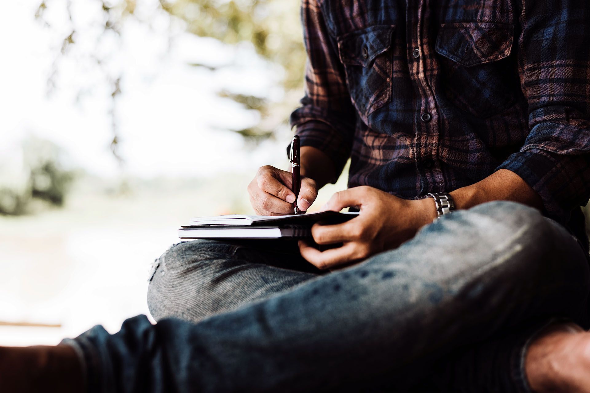 Person sitting cross-legged, writing in a notebook. Wearing a plaid shirt and jeans. Outdoors, writing.