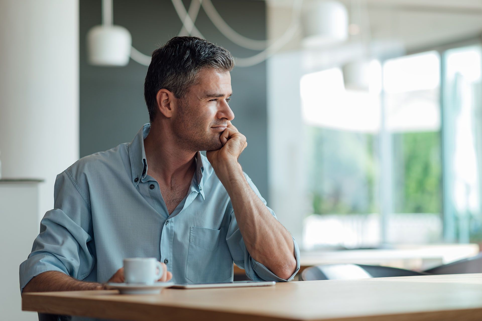 Man sits at a table, resting chin on hand and looking to the side. Coffee cup present. Bright setting.