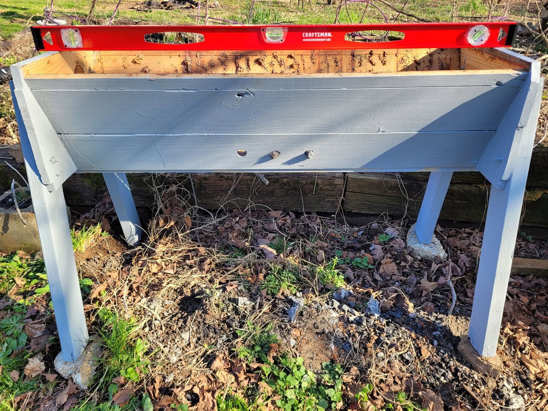 Gray wooden planter box with a red level resting on top outdoors over dirt and leaves