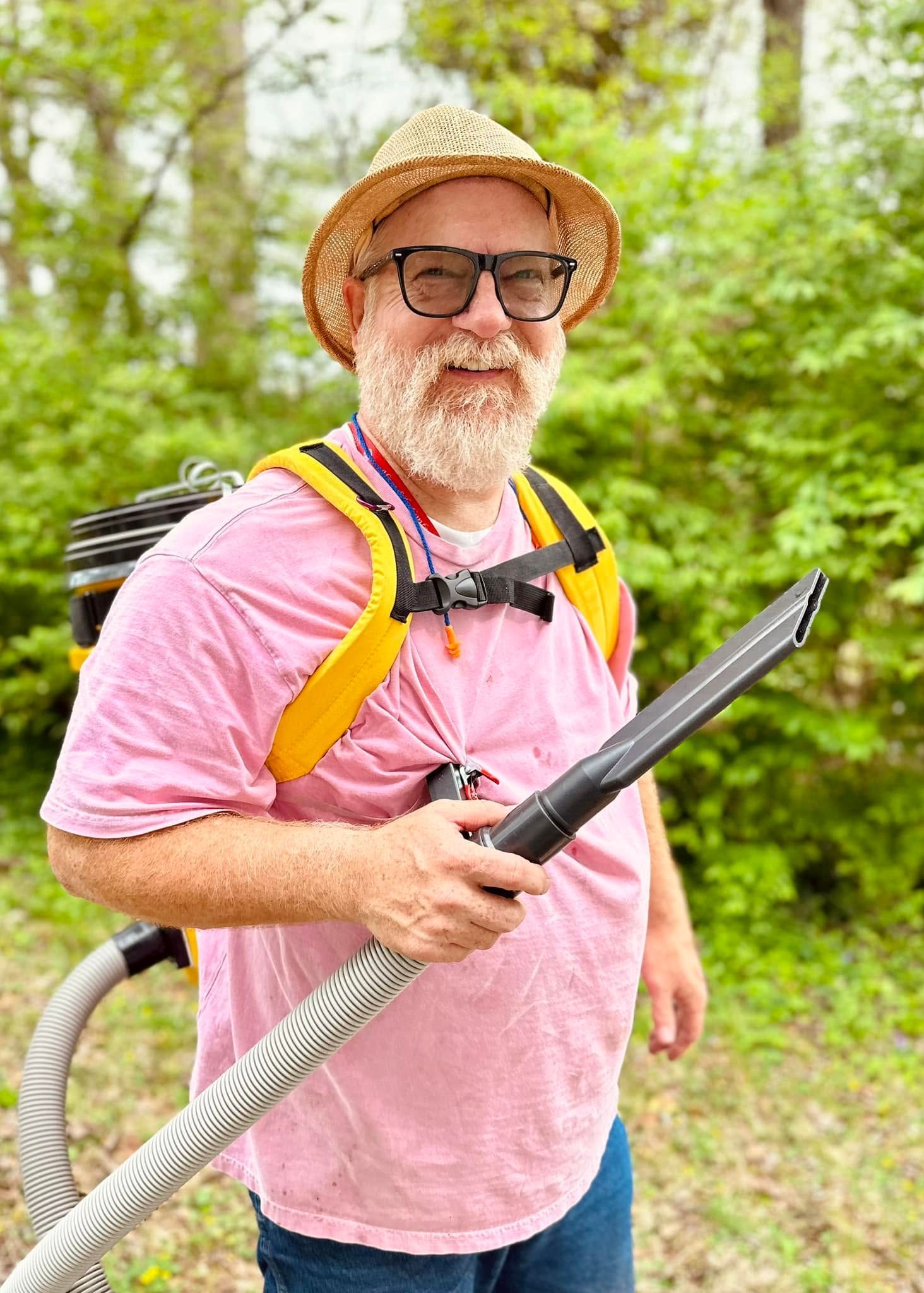 Man in pink shirt and straw hat using a leaf blower in a grassy wooded area