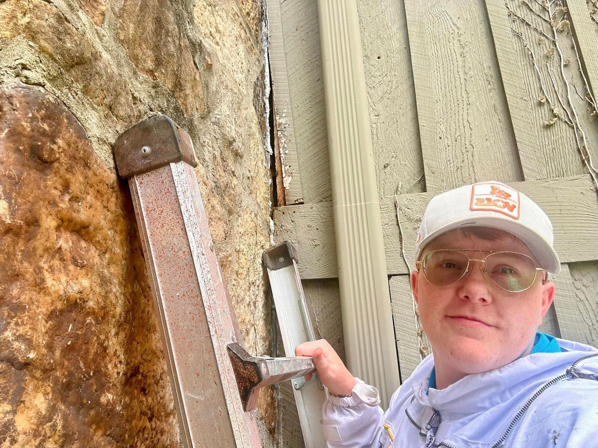 Person in cap and glasses holding a tool next to a rock wall and wooden siding outdoors.