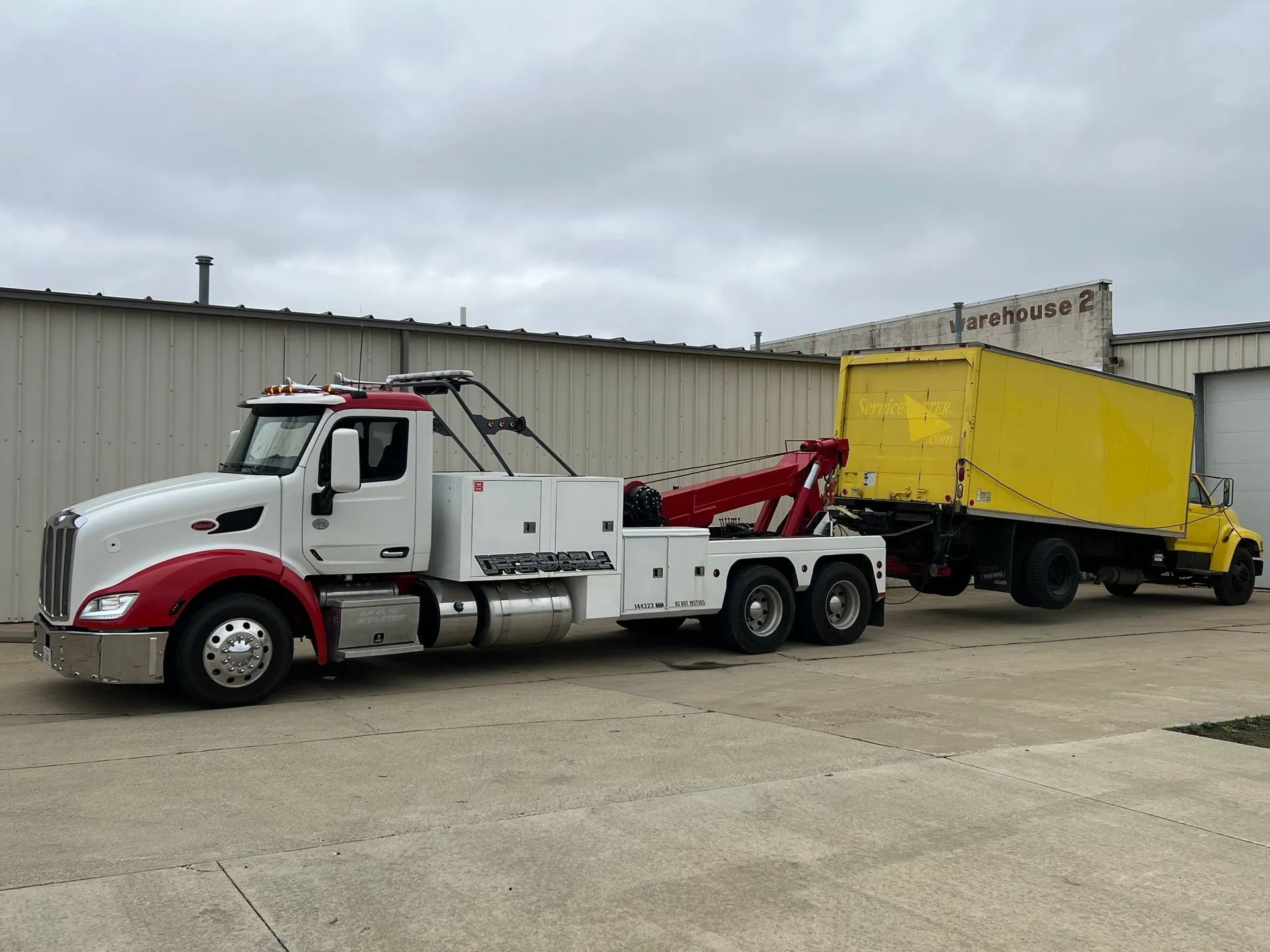 White tow truck hauling a yellow trailer in an industrial lot under cloudy skies