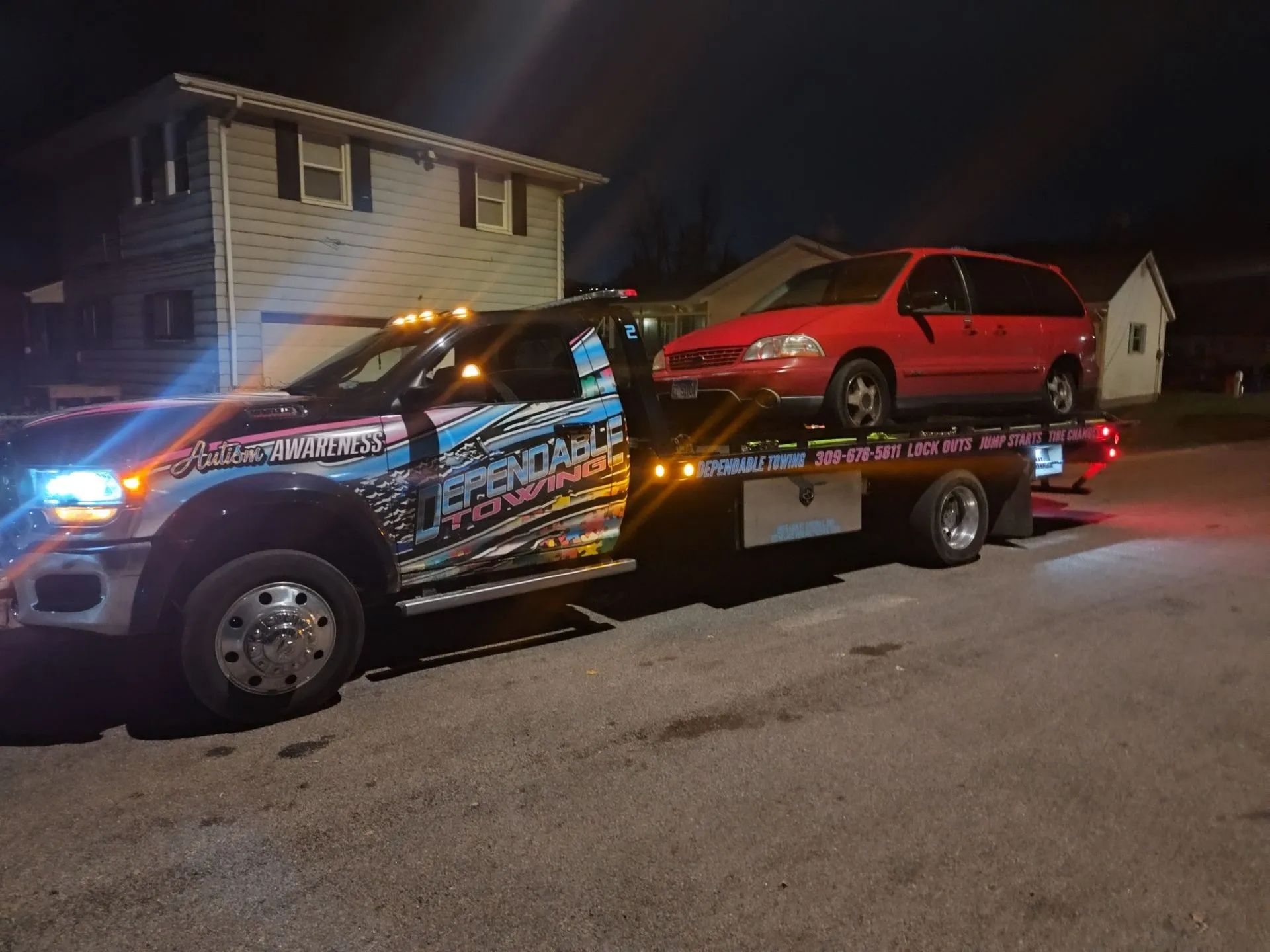 Tow truck carrying a red minivan at night. The truck has colorful decals; a house is in the background.
