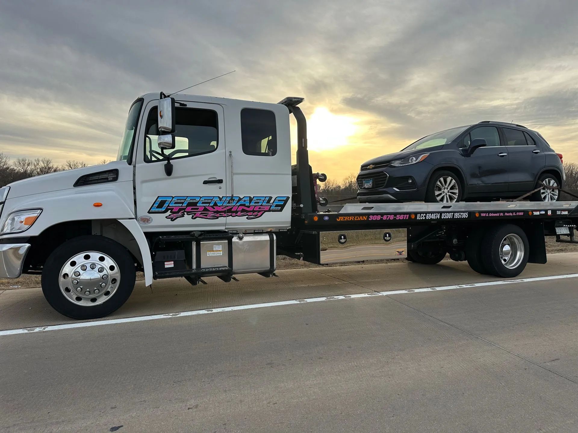A white flatbed tow truck carrying a dark SUV parked on a paved road during a sunset.