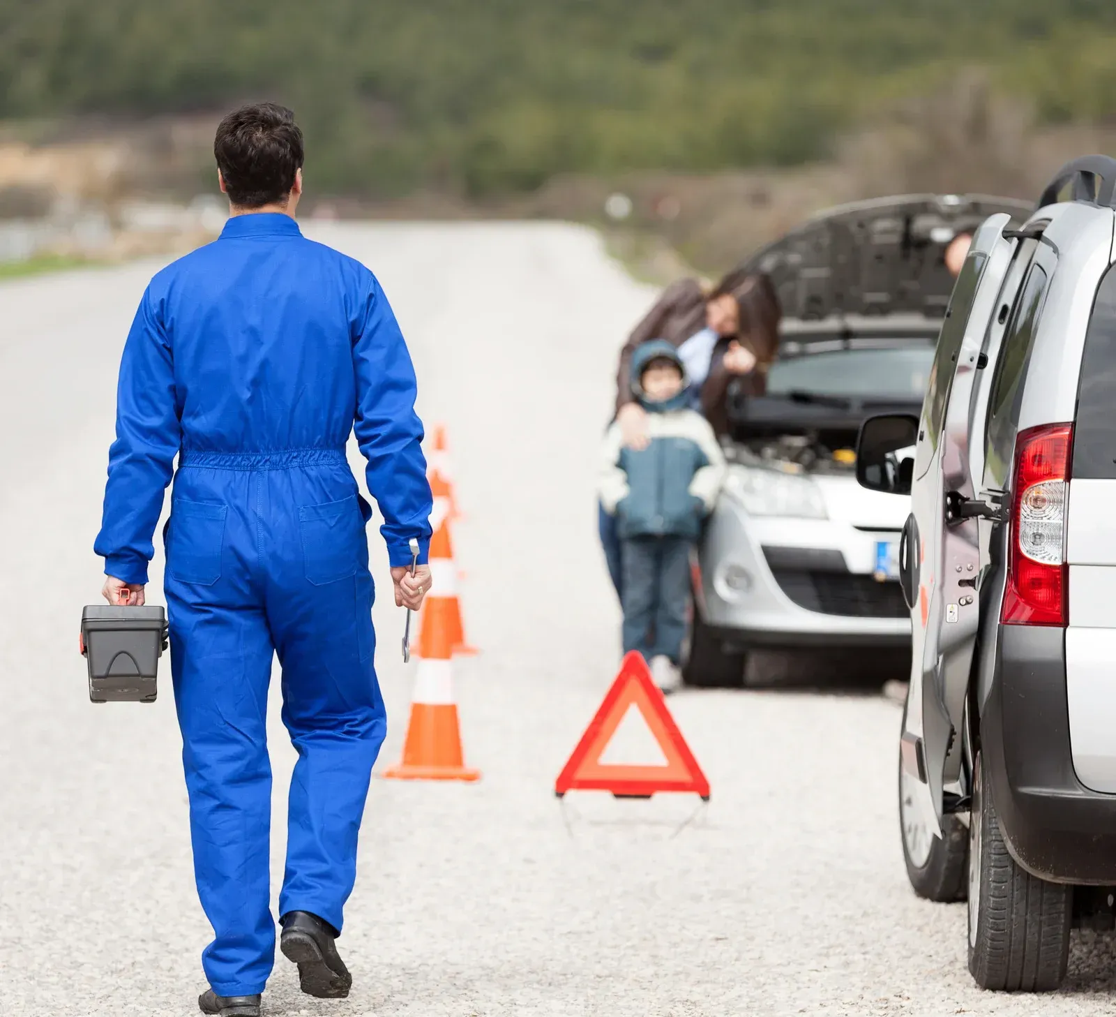 Mechanic walking toward a car with a family, roadside assistance. Red warning triangle, cones. Blue coveralls, toolbox.