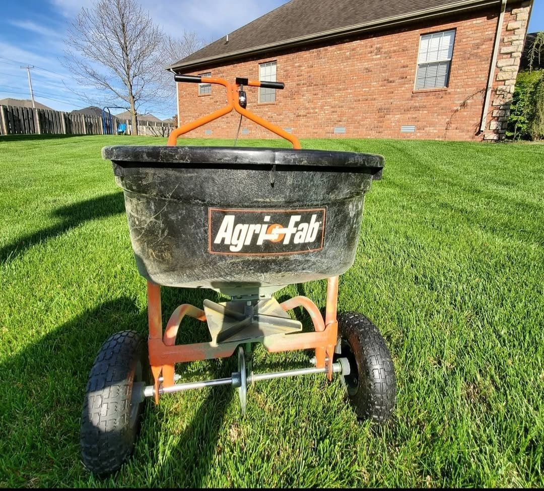 A lawn spreader is sitting on top of a lush green lawn in front of a brick house.
