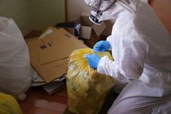 Person in protective suit, gloves, and respirator seals a yellow trash bag in a cluttered room.