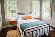Bedroom with iron bed frame, white bedding, teal blanket, wood nightstand, and three windows.