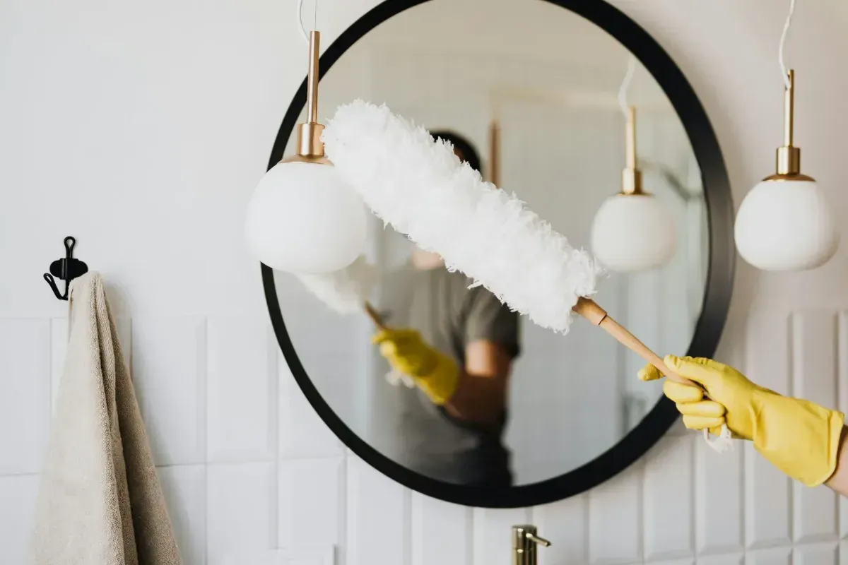 A person wearing yellow gloves dusts a light fixture in a bathroom with a round mirror.