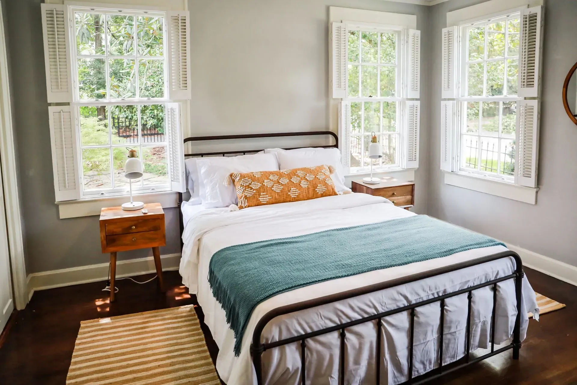 Bedroom with iron bed frame, white bedding, teal blanket, wood nightstand, and three windows.