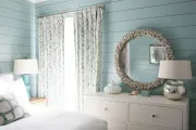 Bedroom with blue shiplap walls, white dresser, large shell-framed mirror, and patterned curtains.