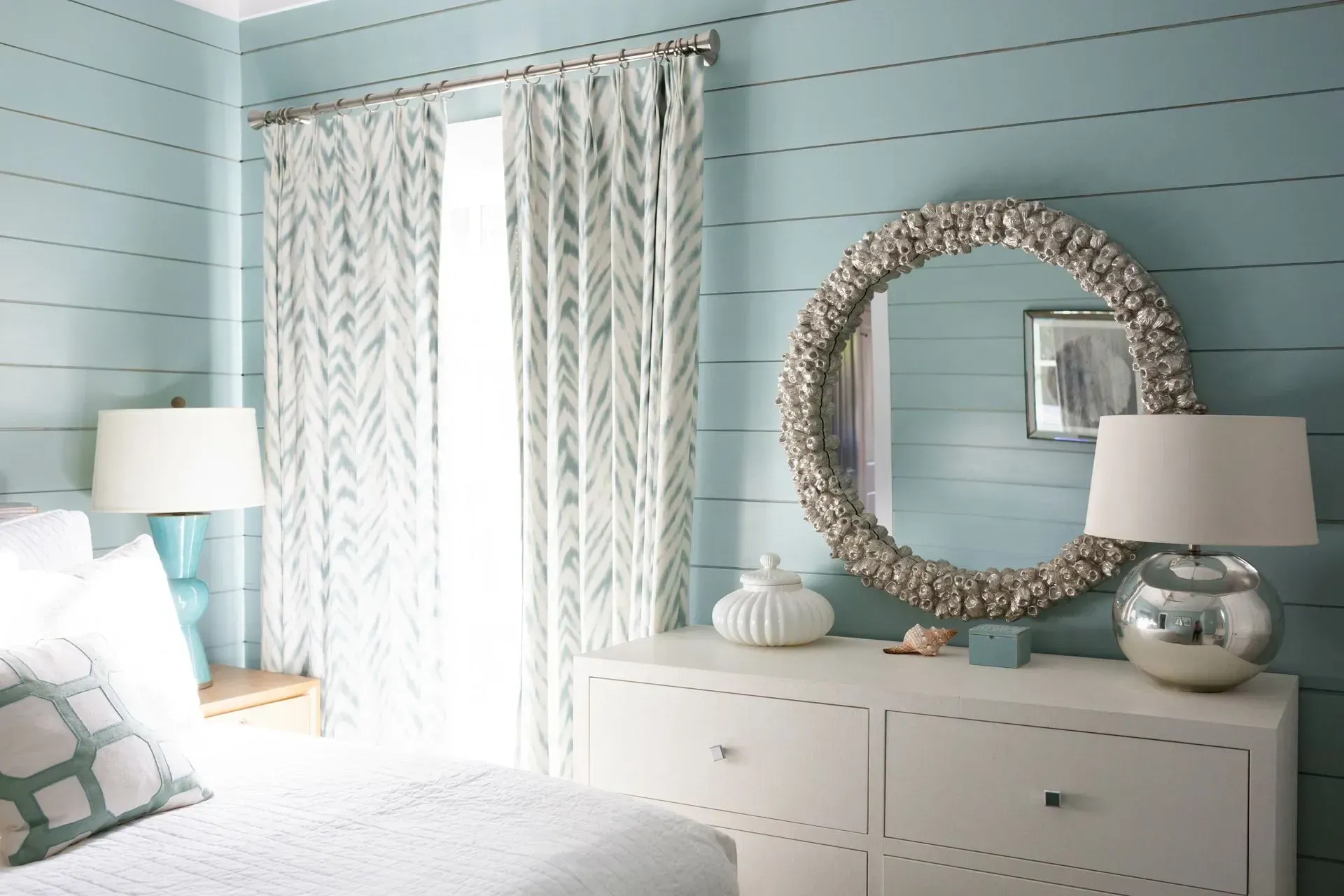 Bedroom with blue shiplap walls, white dresser, large shell-framed mirror, and patterned curtains.