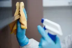 Person in blue gloves spraying cleaner onto a surface and wiping with a yellow cloth.