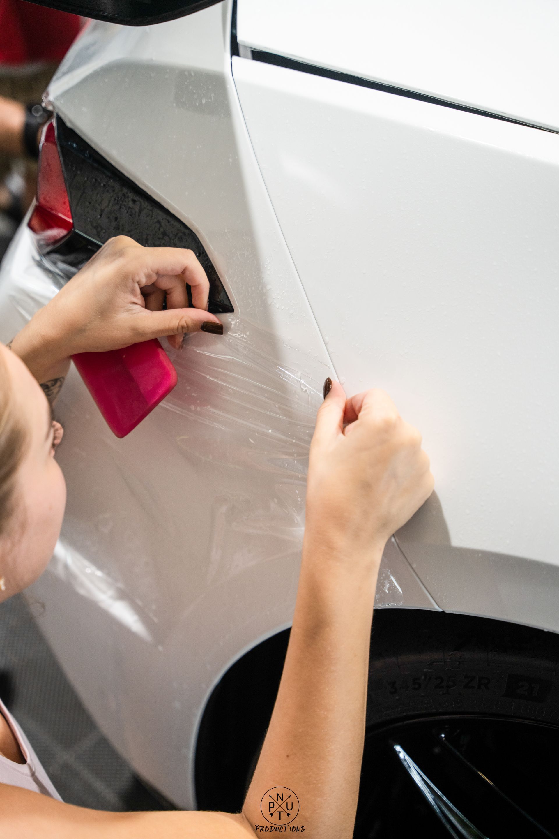 Hands applying clear protective film to a white car door panel near the rear wheel.