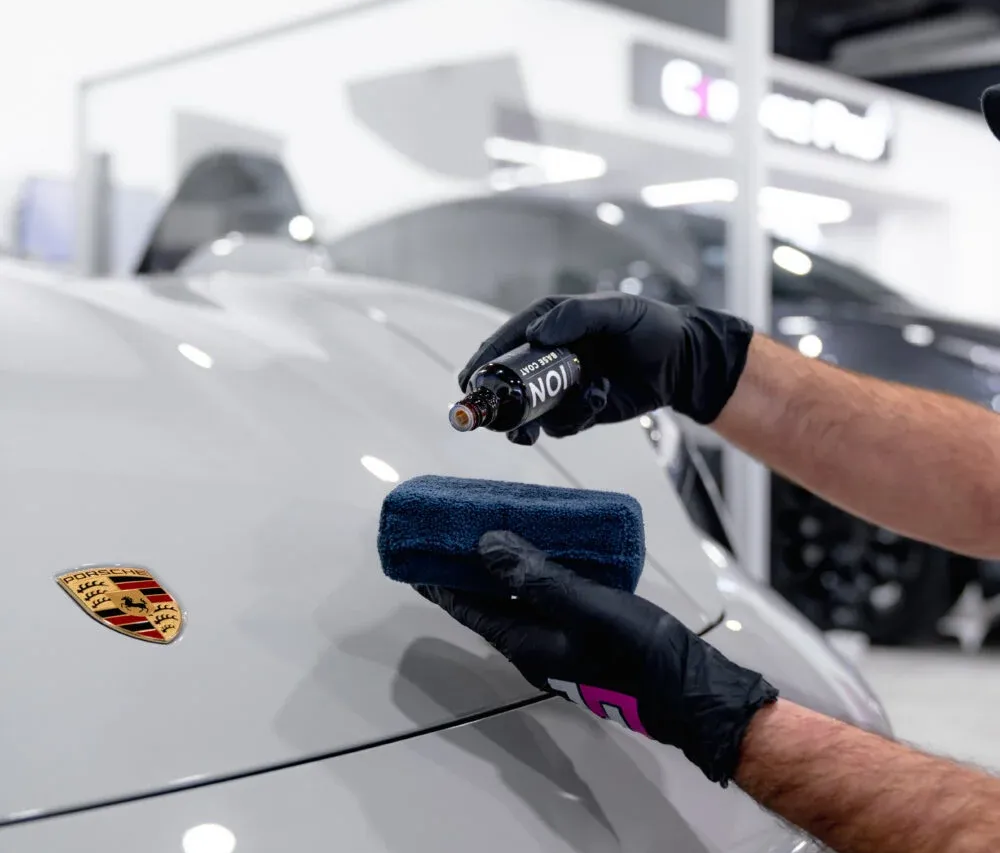 Gloved hands polishing a white Porsche hood with a microfiber cloth and spray in an auto shop