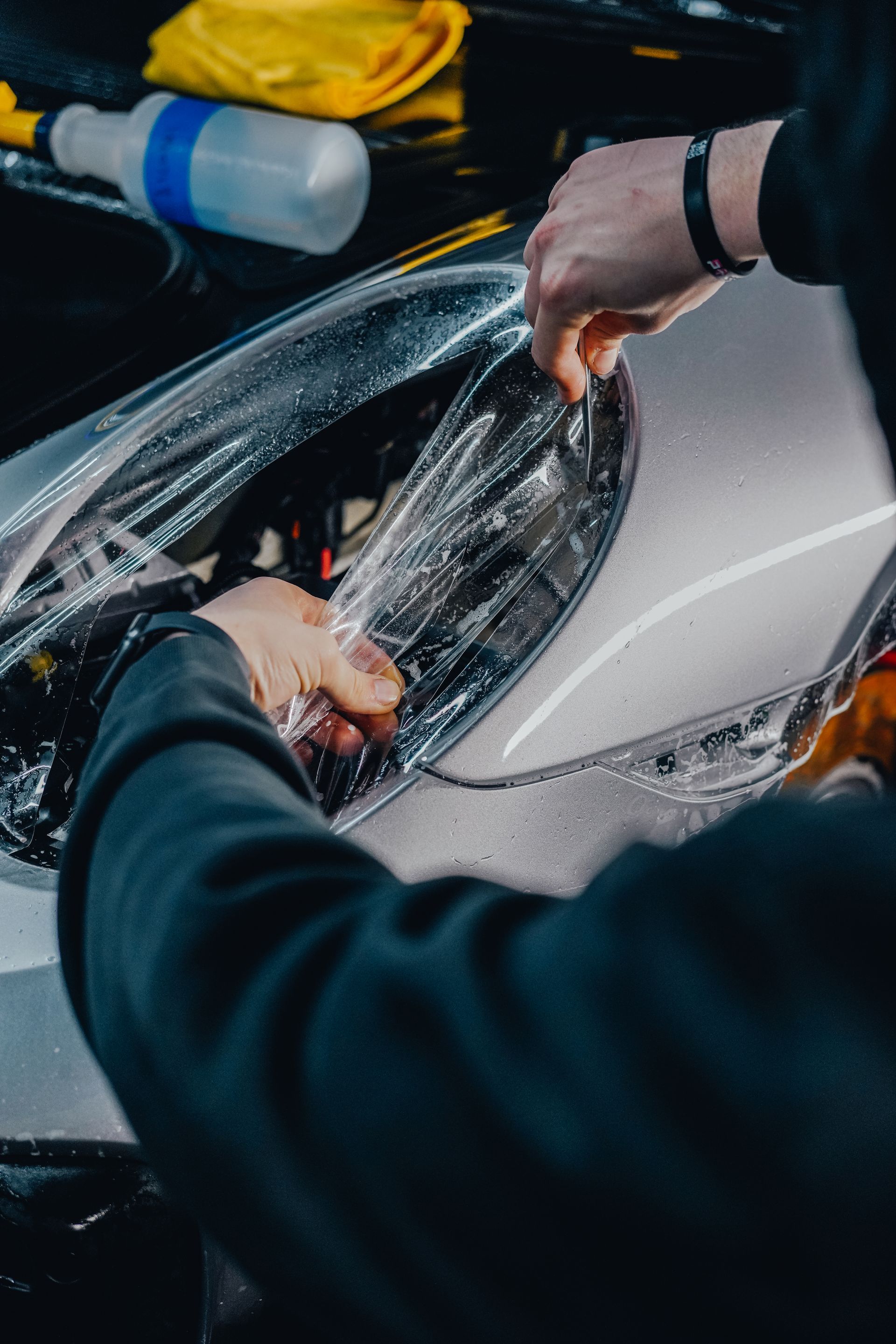 Hands installing clear protective film on a white car headlight with a spray bottle nearby