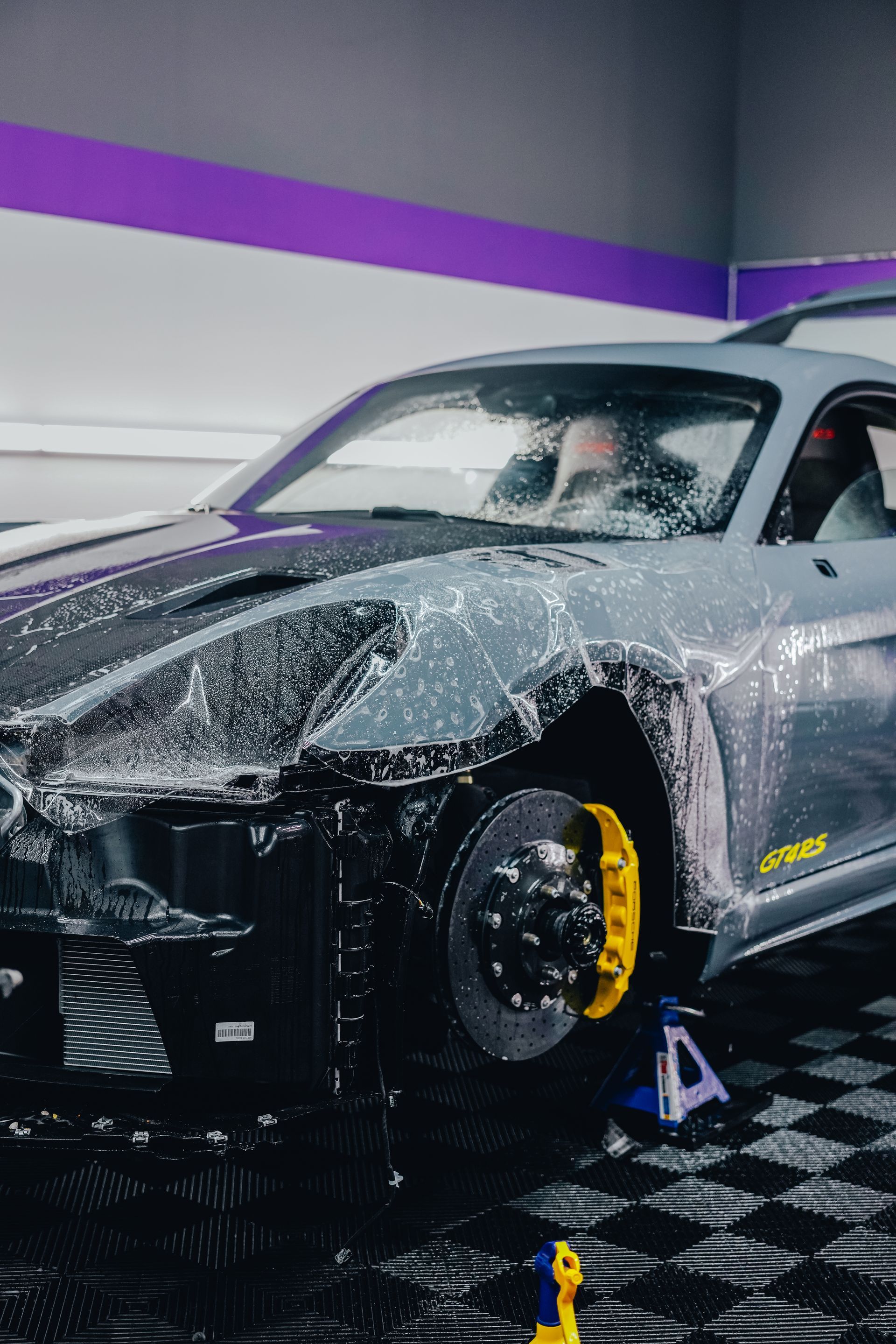 Sports car being sprayed with water in an indoor detailing bay, with yellow wheel covers and white foam.