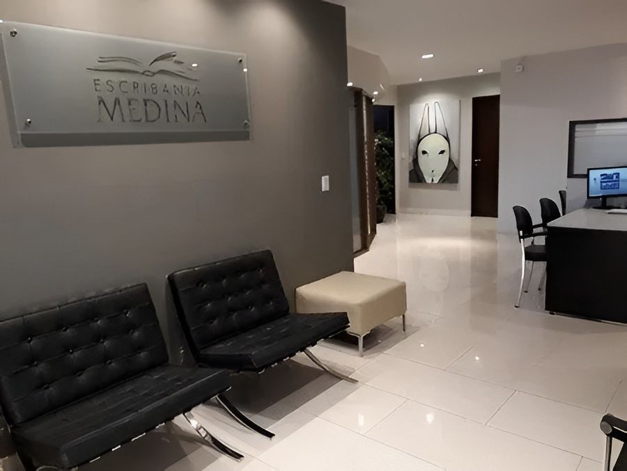 Vestíbulo de clínica moderna con sillas negras, banco beige y mostrador de recepción en un pasillo luminoso.