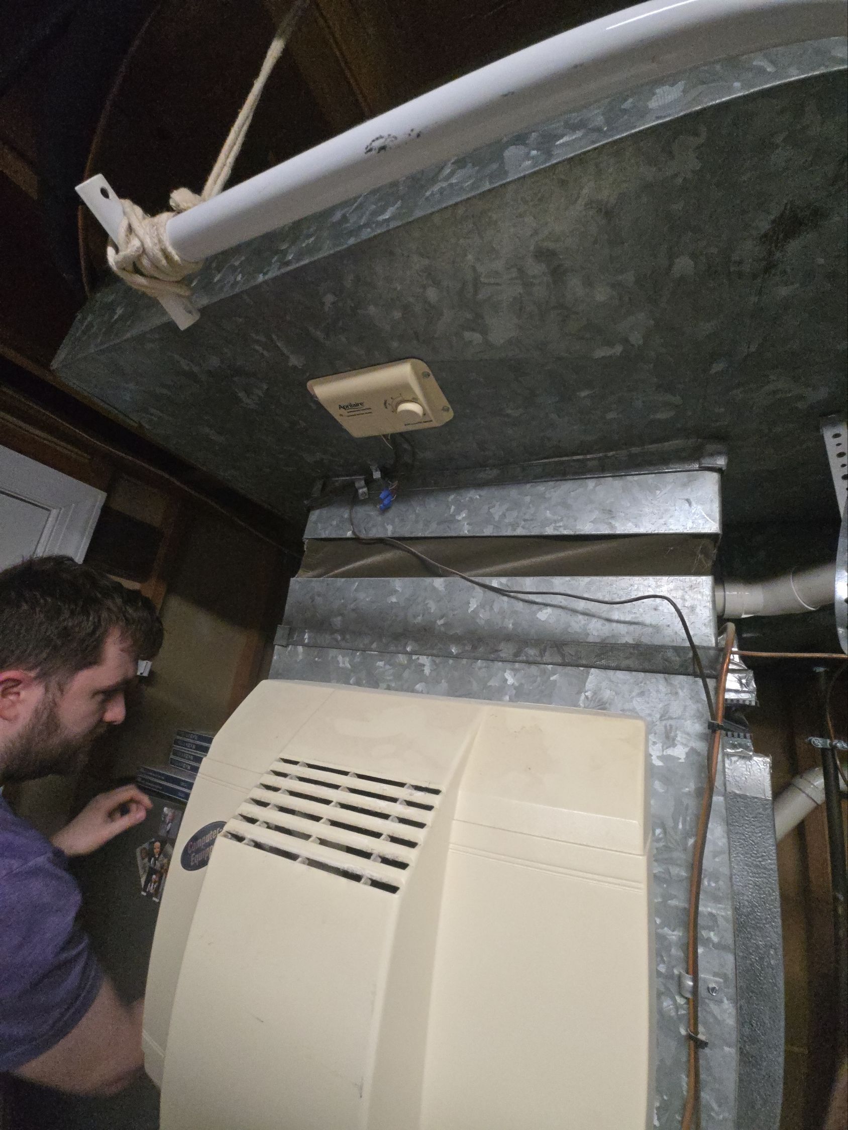 A person in a purple shirt inspects the metal ductwork and control panel of a residential HVAC unit in an attic.