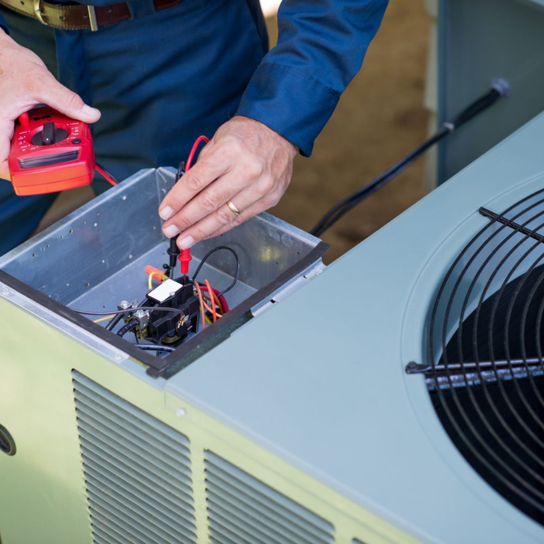 A technician uses a red multimeter to test the electrical components inside an open outdoor air conditioning unit.