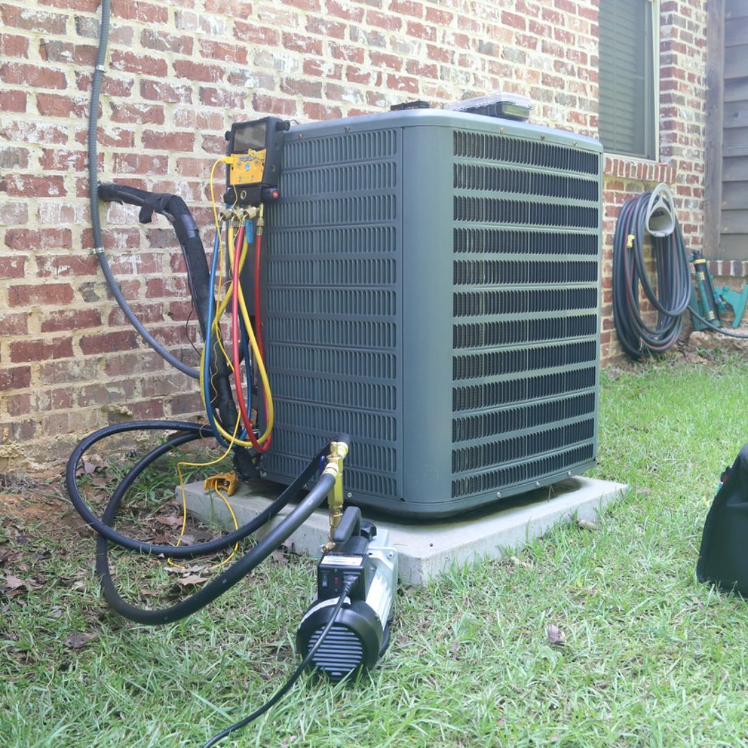 An HVAC unit outside a brick house connected to a vacuum pump and gauge manifold for maintenance.