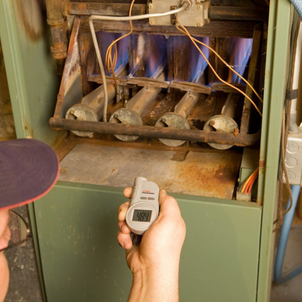 A technician holds a digital meter to measure the temperature of the lit burners inside an open residential furnace.