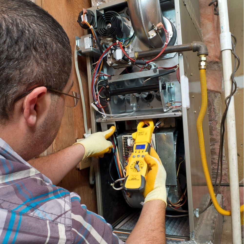A technician in yellow gloves uses a multimeter to inspect the wiring inside an open residential furnace.