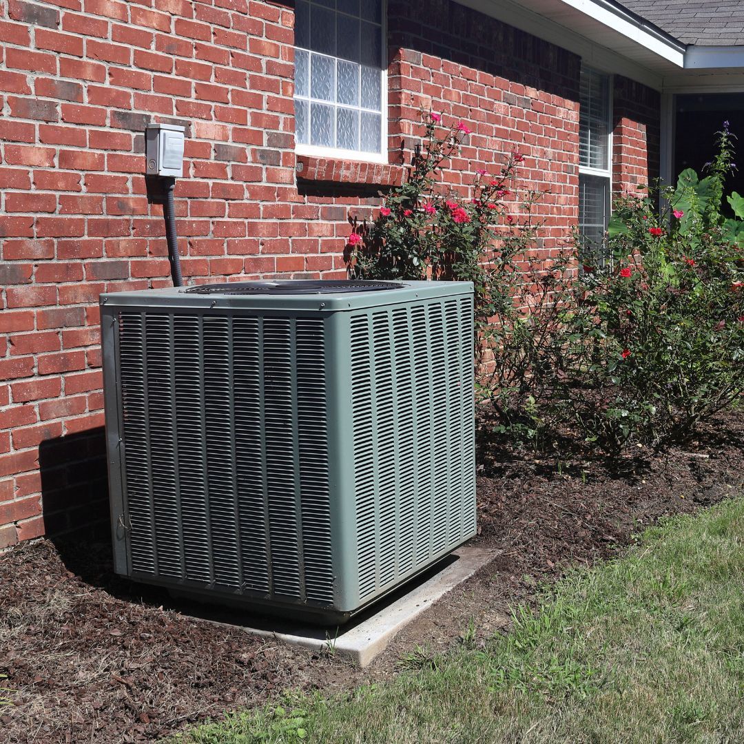 A grey HVAC condenser unit sits on a concrete pad next to a brick house wall surrounded by garden bushes.