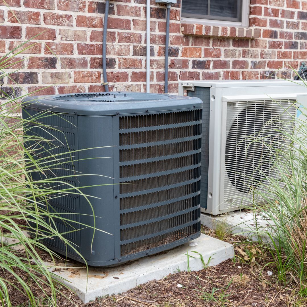 Two outdoor air conditioning units sit on concrete pads against a brick exterior wall near ornamental grasses.