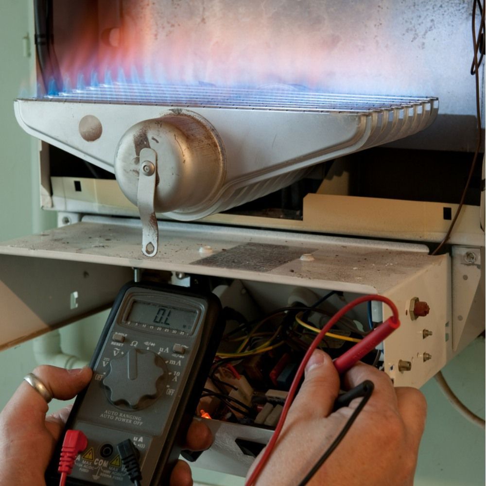 A person uses a multimeter to test internal electrical components of an active, blue-flamed gas boiler.