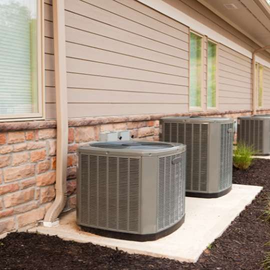 Three beige HVAC outdoor condenser units on a concrete pad next to a building with stone siding and horizontal tan panels.
