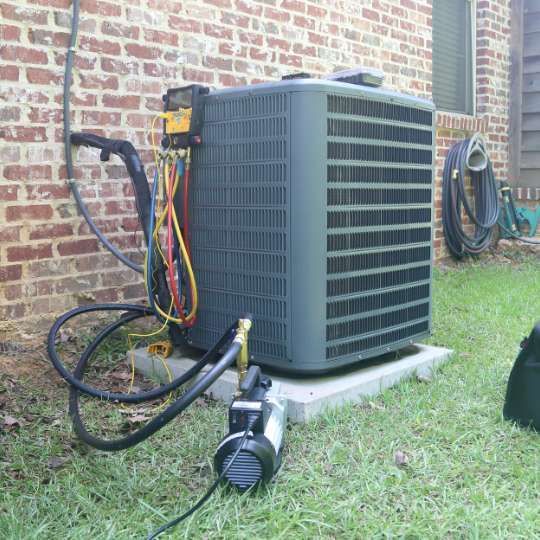 A vacuum pump connected to an outdoor HVAC unit via a manifold gauge set sitting on a concrete pad by a brick wall.