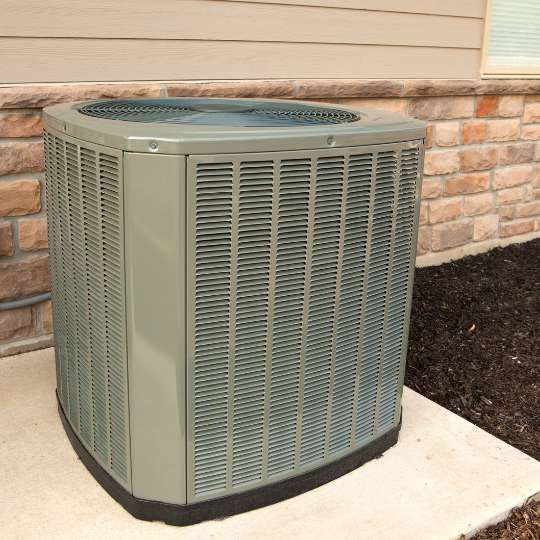 A beige, square central air conditioning unit sits on a concrete pad against a house with stone siding.