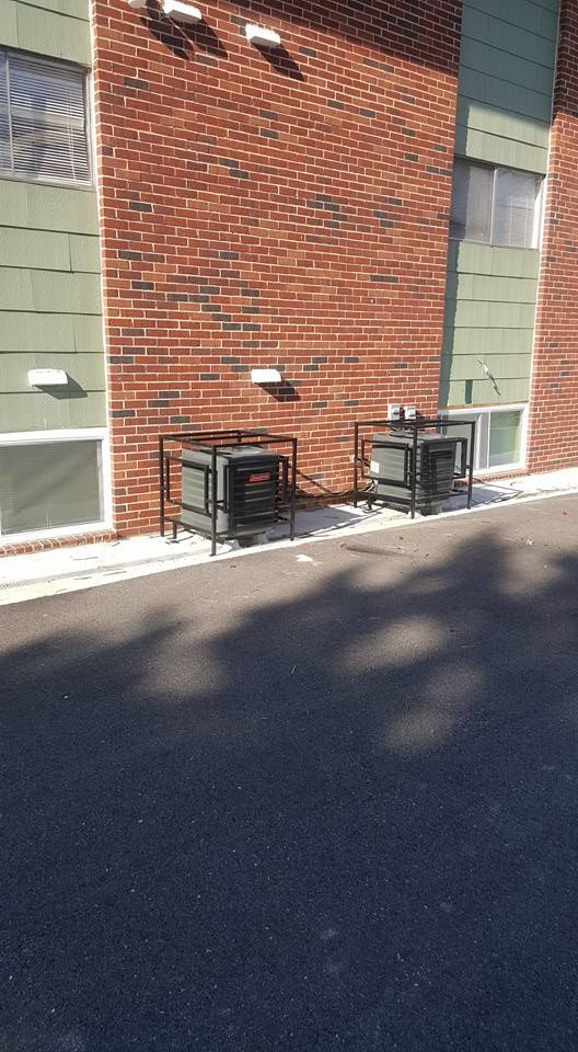 Two outdoor HVAC units sitting on a concrete pad against a red brick building with green siding and basement windows.