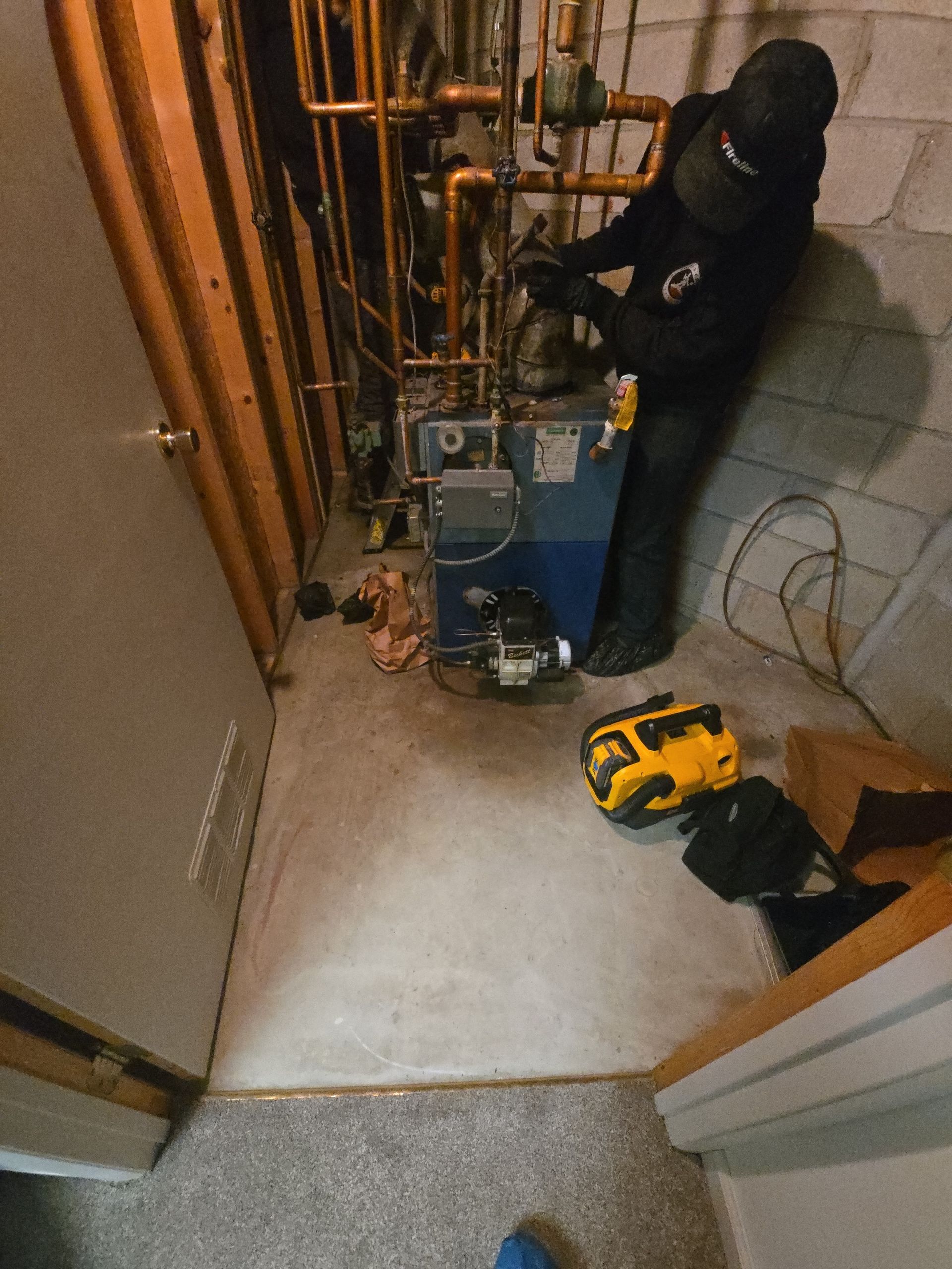 A technician in black work clothes services a boiler in a utility room with exposed copper pipes and concrete walls.
