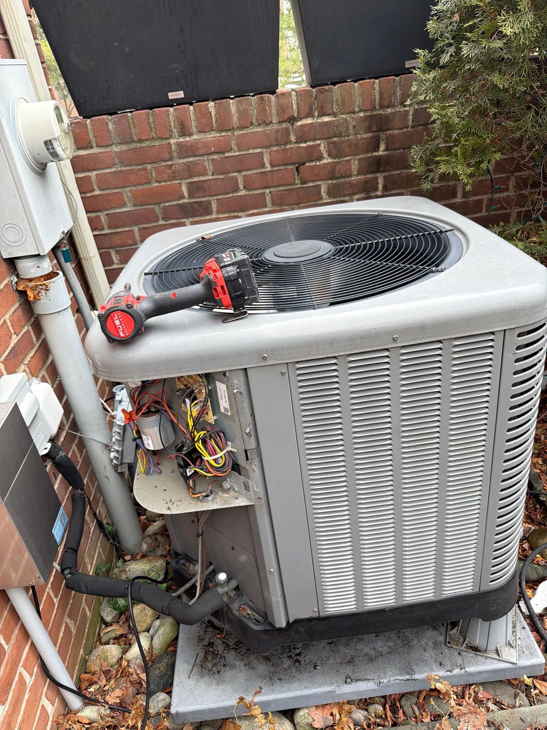 Outdoor HVAC unit with a red-handled tool resting on top, beside a brick wall and utility lines