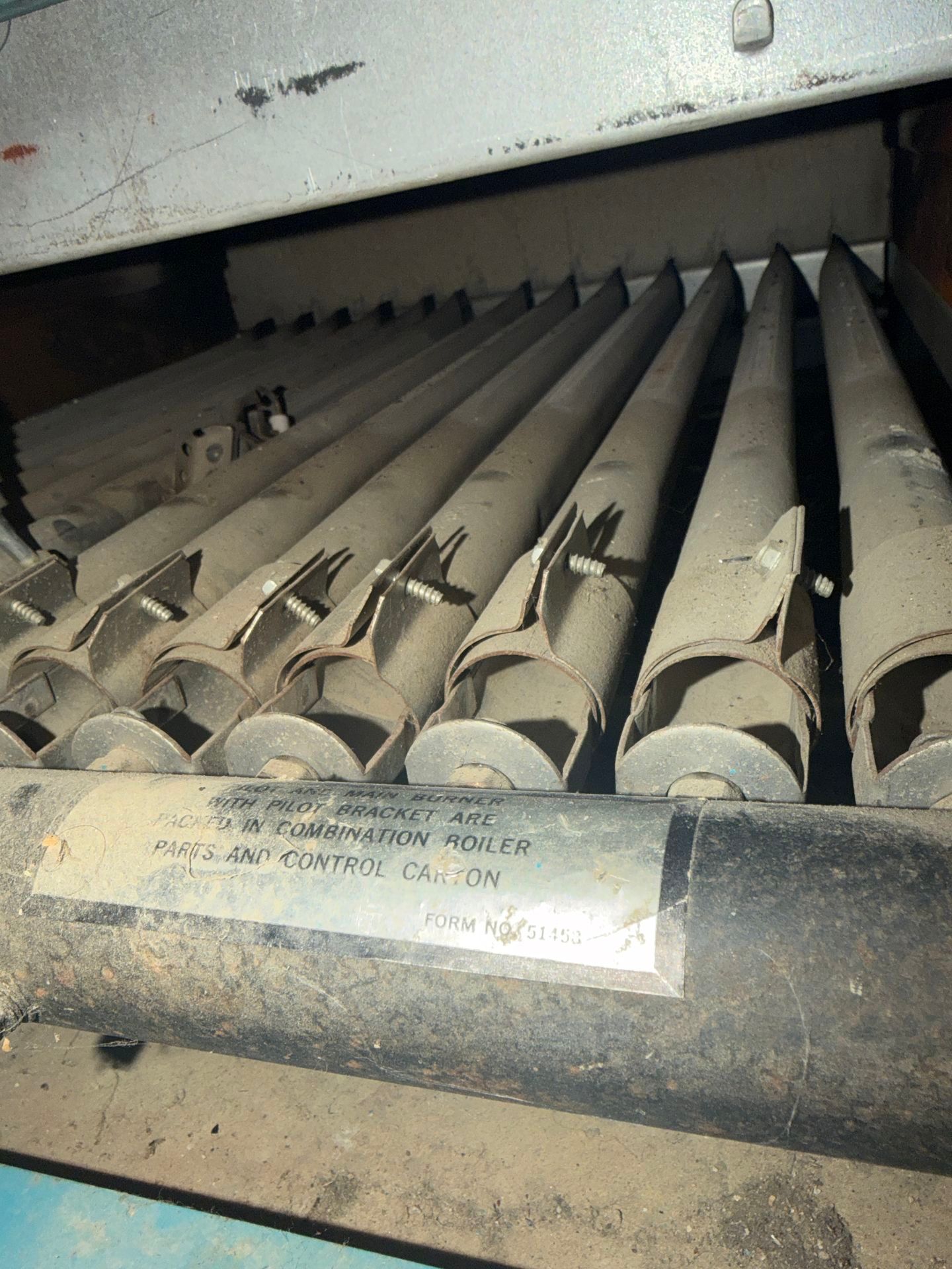 A close-up of several parallel burner tubes inside a dusty furnace, with a faded warning label on the front manifold.