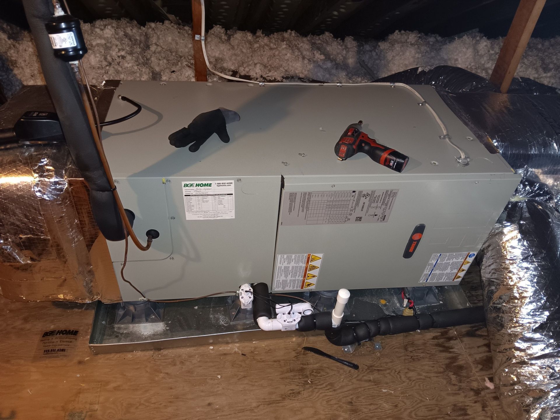 An HVAC air handler unit sitting on a metal drain pan in an attic with tools placed on top of it.