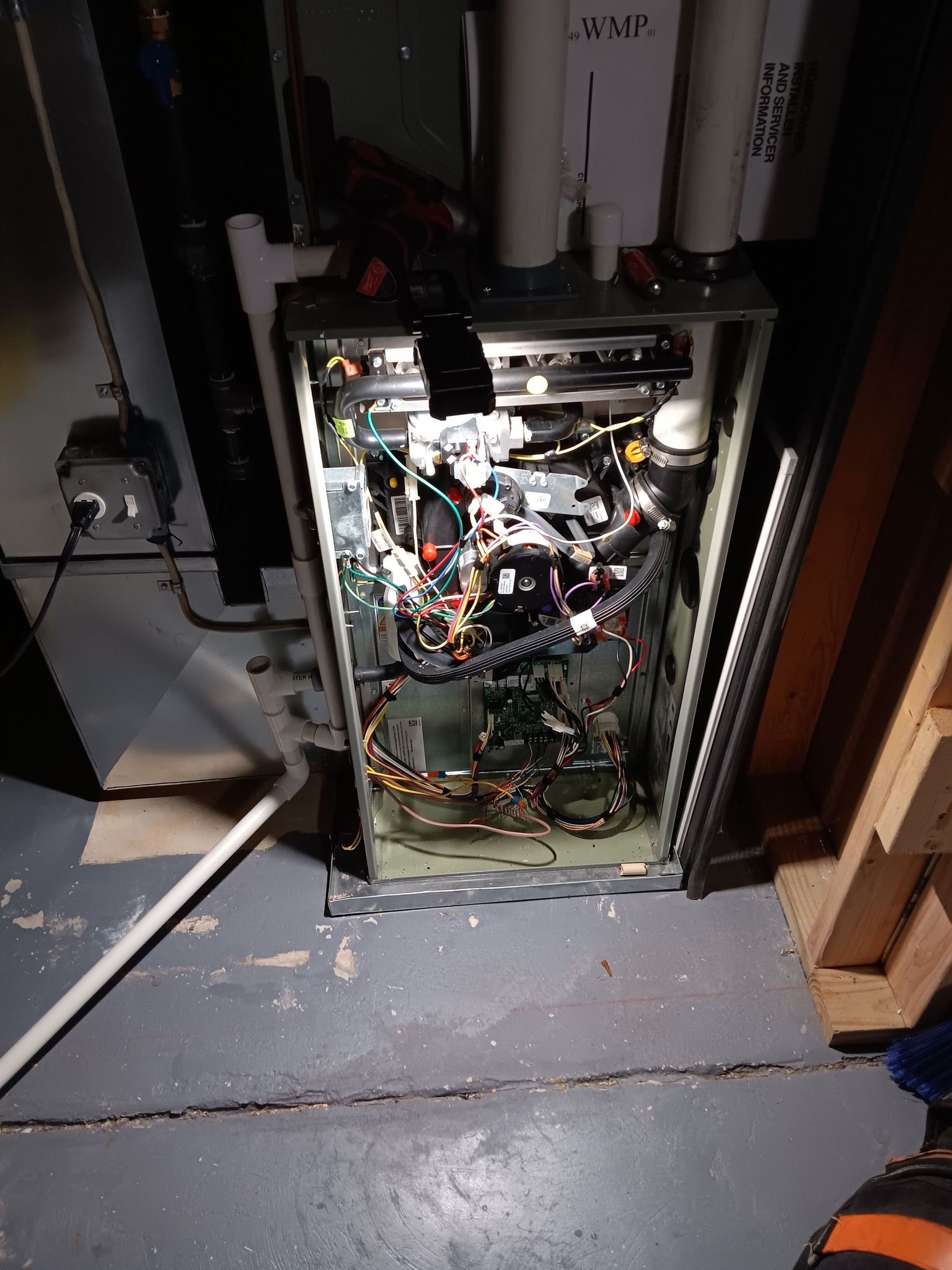 An open furnace interior in a basement setting, revealing complex wiring, components, and metal piping.