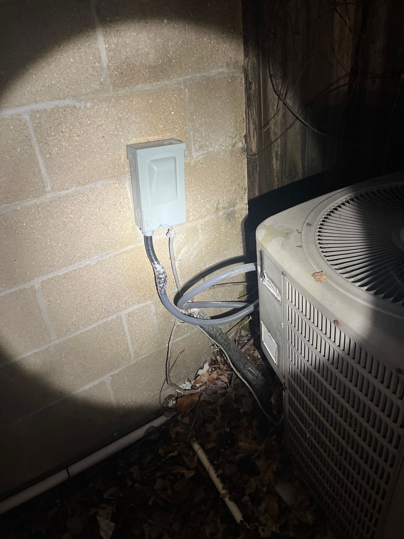 An outdoor air conditioning unit sits next to a cinderblock wall with a grey electrical disconnect box mounted above it.