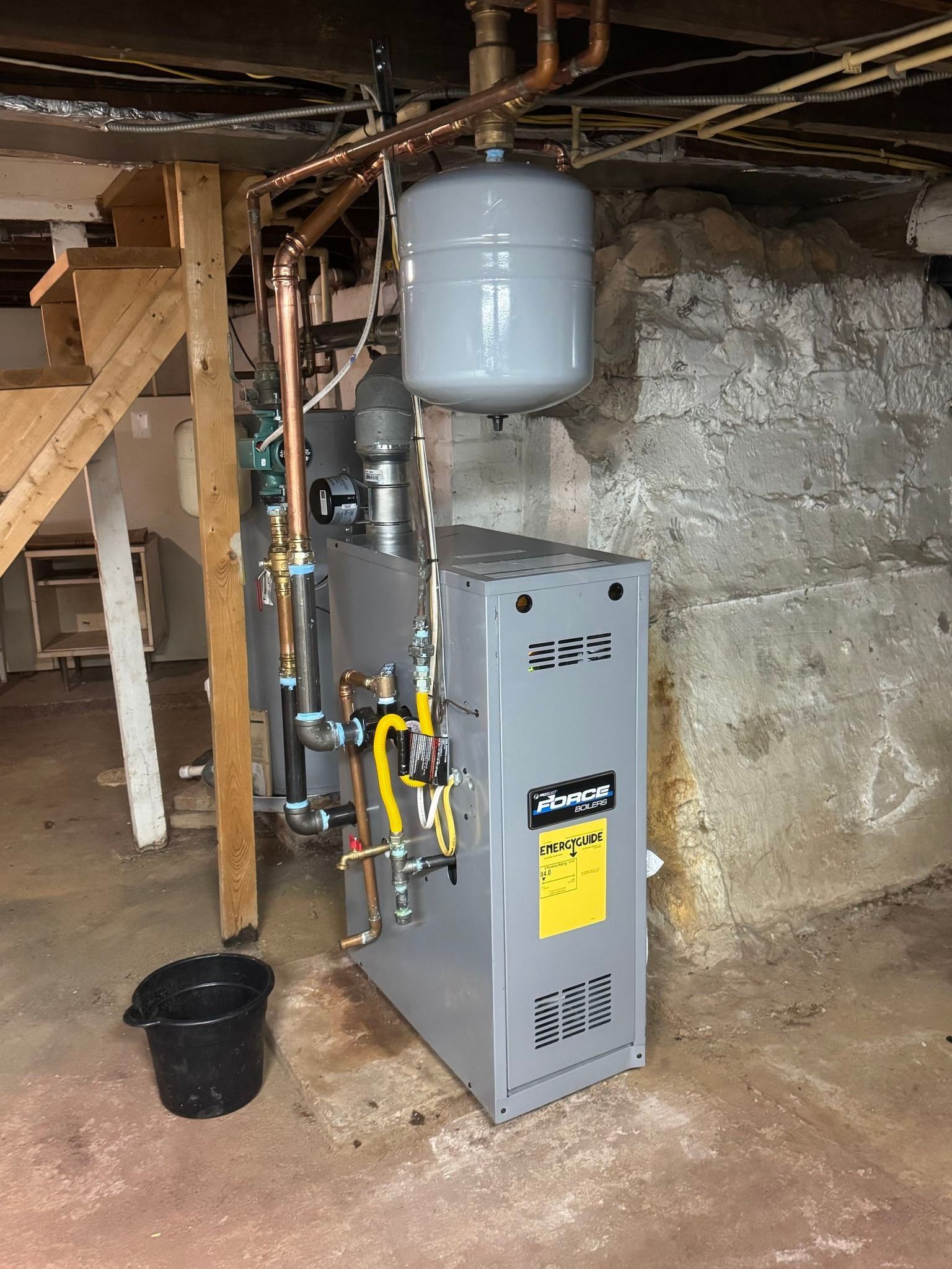 A grey residential boiler system with a connected expansion tank and copper piping in a basement with a stone wall.