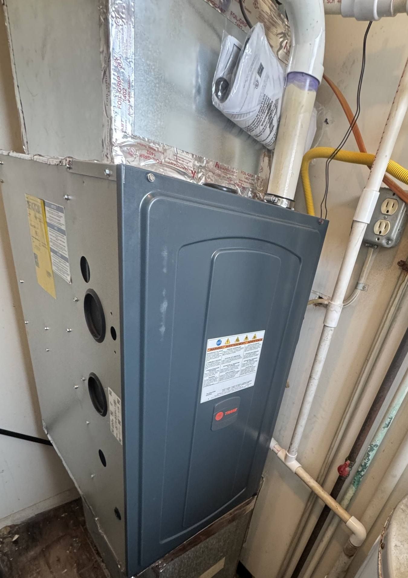 A gray Trane furnace unit installed in a utility closet with connected white PVC venting pipes and metallic ductwork.