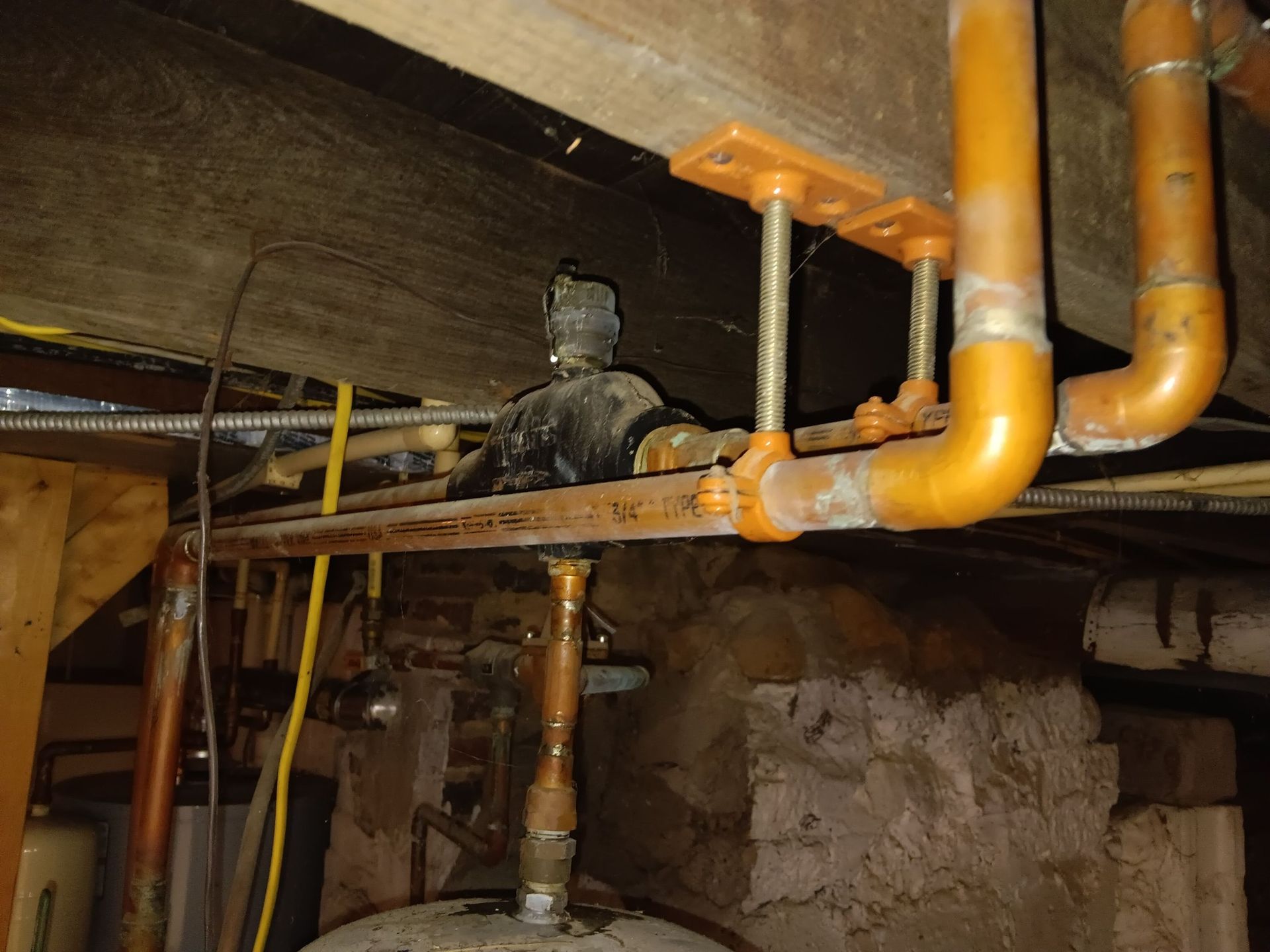 Copper plumbing pipes with hangers attached to an overhead wooden floor joist in a basement or crawl space.