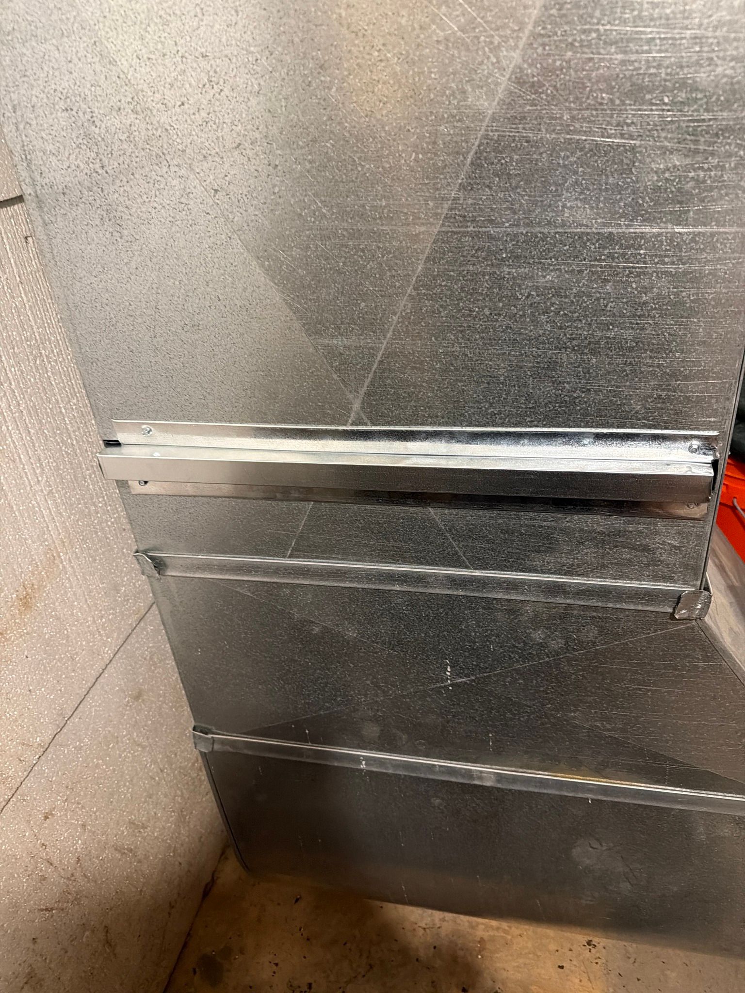 A close-up of galvanized metal HVAC ductwork showing a horizontal seam connection against a concrete wall.