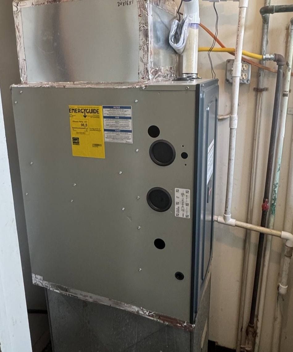 A gray HVAC furnace unit with a yellow energy guide label, installed indoors with nearby utility pipes.